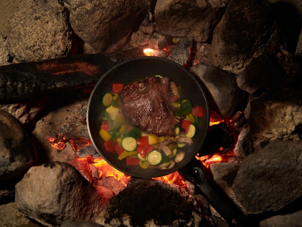 Steak and vegetables cooking in a cast iron skillet over campfire coals